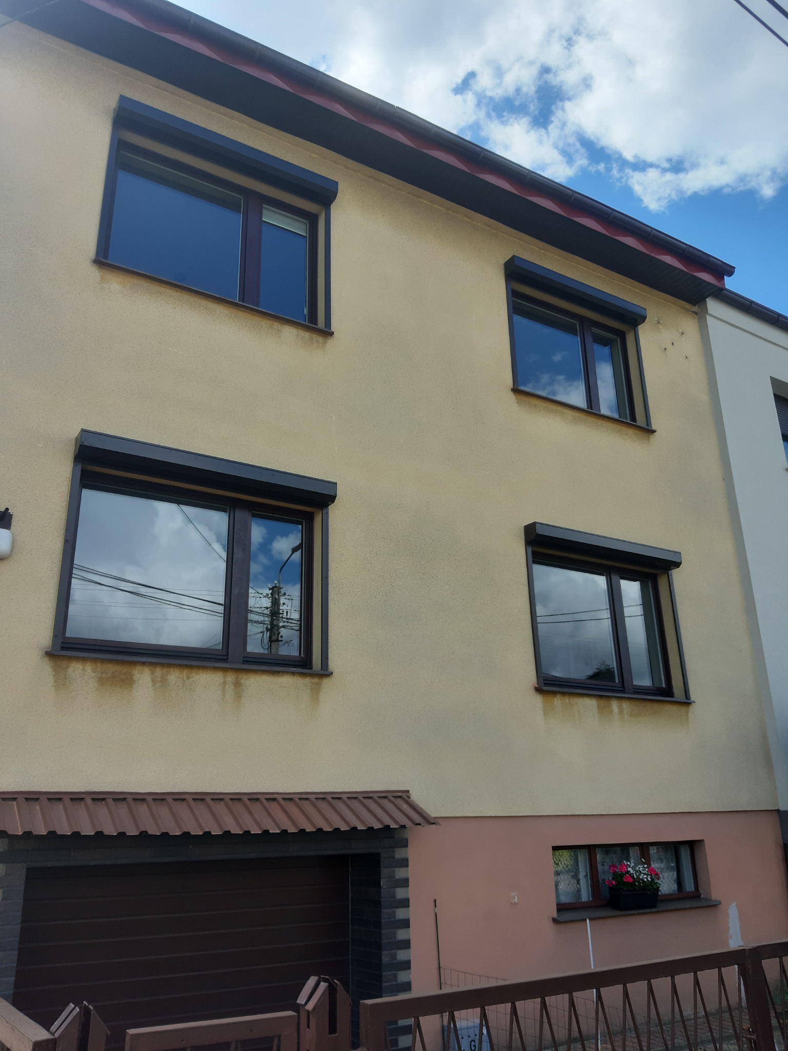 Dom z oknami PCV i roletami zewnętrznymi w kolorze grafitowym, odbicie chmur w szybach, widoczna brama garażowa i okno z kwiatami na parterze.