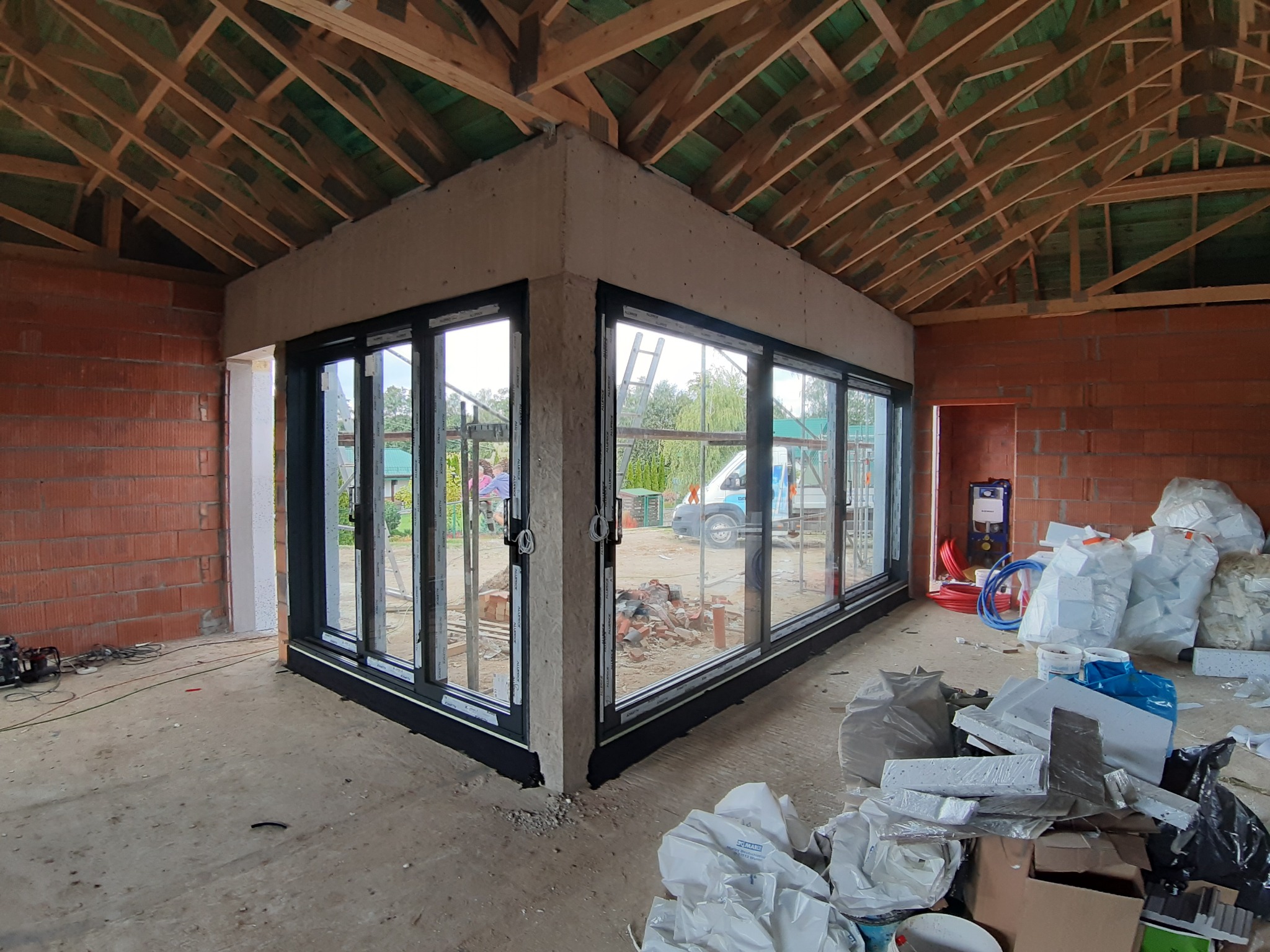 Wnętrze w trakcie budowy z zamontowanymi przesuwnymi oknami z czarnymi ramami, widok na zewnątrz z zielenią i zaparkowanym białym busem, surowy stan wykończenia ścian z cegły i drewnianej...