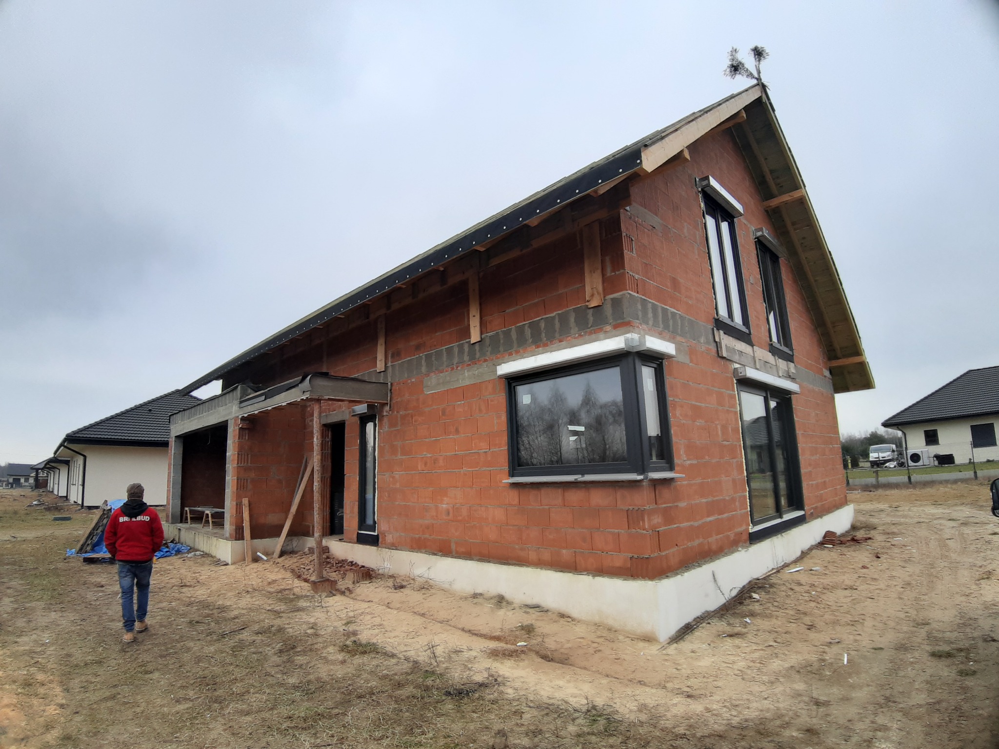 Surowy budynek jednorodzinny w trakcie budowy, widoczne czarne ramy okienne, ściany z czerwonej cegły dziurawki, widoczny fragment dachu oraz mężczyzna w czerwonej bluzie idący w stronę budynku.