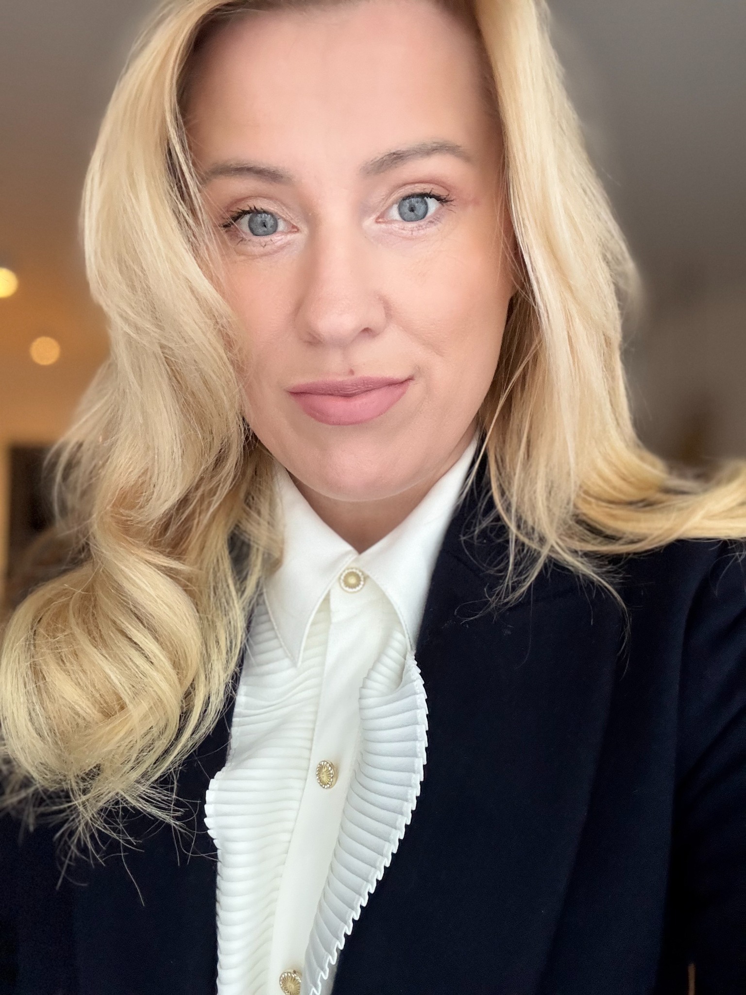 Portret uśmiechniętej blondynki w białej koszuli z plisami i złotymi guzikami oraz w granatowej marynarce, patrzącej w obiektyw.