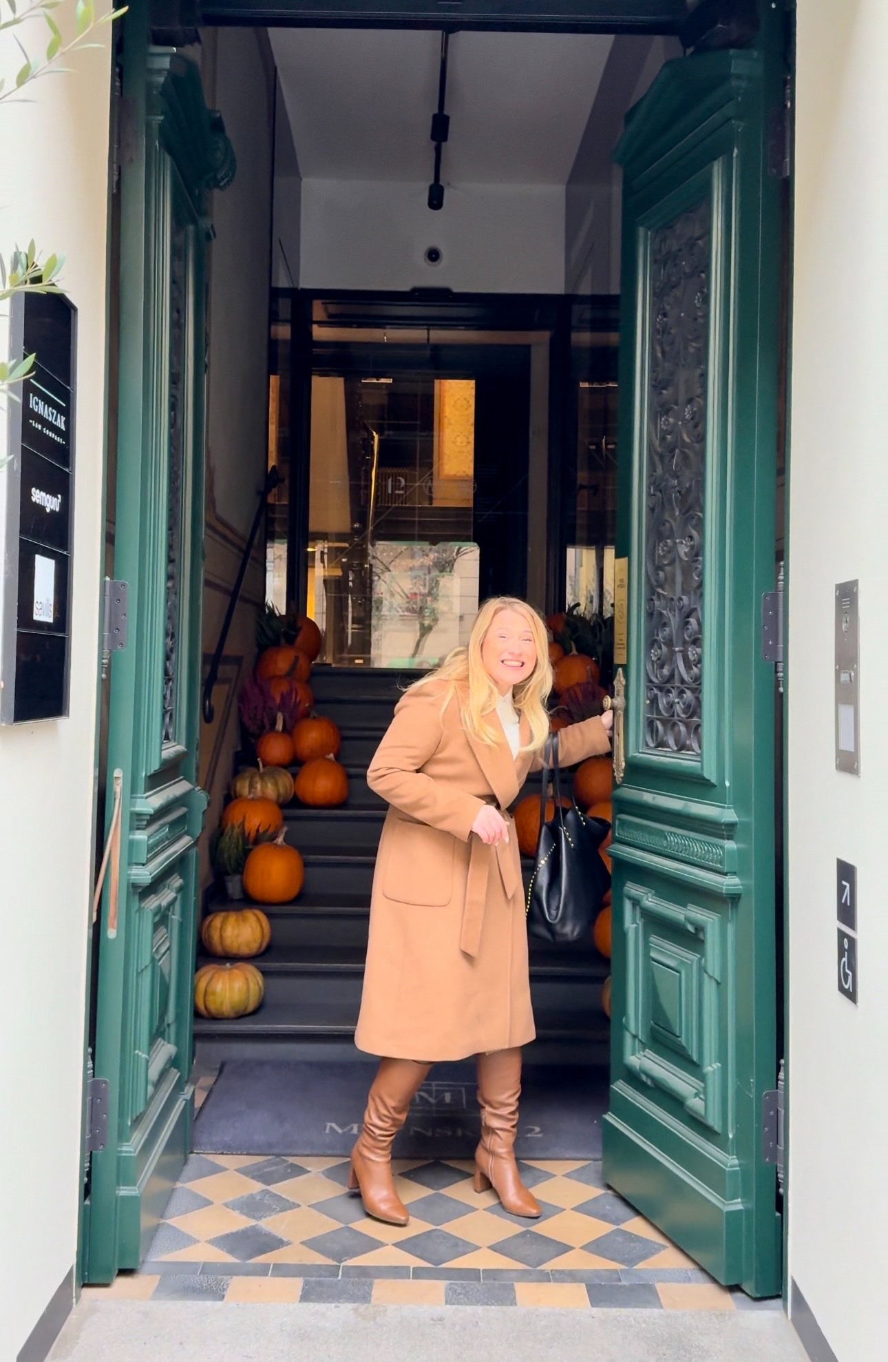 Uśmiechnięta kobieta w płaszczu i kozakach wchodzi do budynku przez szerokie, zielone drzwi. Schody ozdobione dyniami.