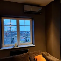 Czarny klimatyzator ścienny z wyświetlaczem temperatury w nowoczesnym salonie z oknem na miasto i brązowymi ścianami, widok z poziomu kanapy.