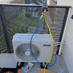 Biała jednostka zewnętrzna klimatyzacji AUX na balkonie, podłączona do manometru cyfrowego za pomocą niebieskiego i żółtego węża, widoczna skrzynka narzędziowa i elementy montażowe.