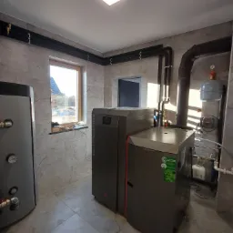 Nowoczesna kotłownia z piecem na pellet i zasobnikiem, rury grzewcze poprowadzone wzdłuż ścian wykończonych płytkami imitującymi marmur.