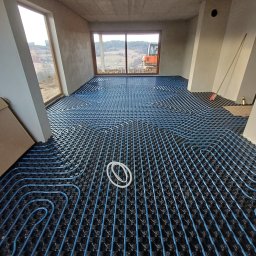 MSJ Instal Marcin Jurkowski - Niebieskie rury ogrzewania podłogowego ułożone na czarnej macie w surowym wnętrzu z oknami wychodzącymi na krajobraz. Widoczny sprzęt budowlany za oknem.