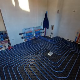 MSJ Instal Marcin Jurkowski - Niebieskie rury ogrzewania podłogowego ułożone na czarnej macie w surowym wnętrzu. Widoczny rozdzielacz i narzędzia. Ujęcie z góry, podkreślające regularny wzór instalacji.