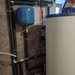 Instalacja grzewcza z niebieskim zbiornikiem wyrównawczym, izolowane rury, biały bojler i karton po akcesoriach w tle, widok z dołu.