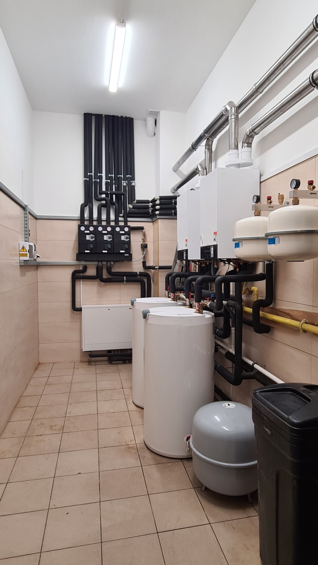 Kotłownia z trzema białymi kotłami, zbiornikami wyrównawczymi i rozbudowaną instalacją rur w izolacji, widoczne także czarne panele sterujące na ścianie z beżowymi płytkami.
