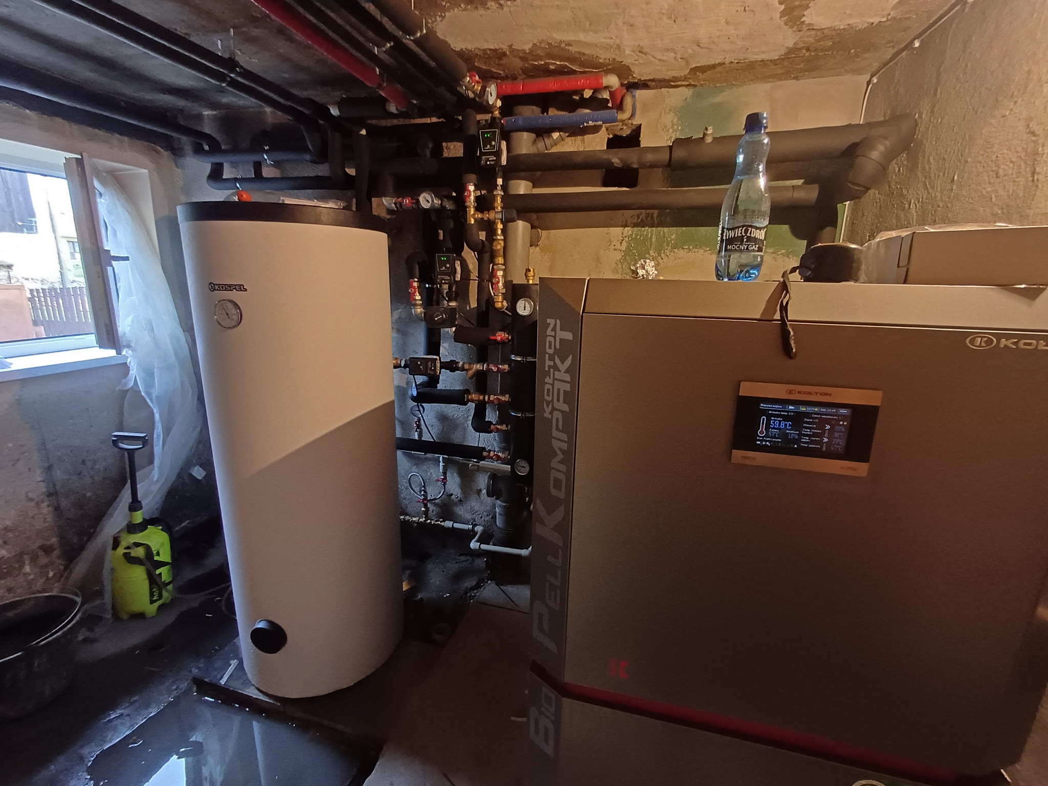 Kotłownia z piecem na pellet Kolton Bio Pellkompakt, zasobnikiem Kospel, licznymi rurami instalacji grzewczej i butelką Żywiec Zdrój na piecu. W tle okno piwniczne i ślady wilgoci.