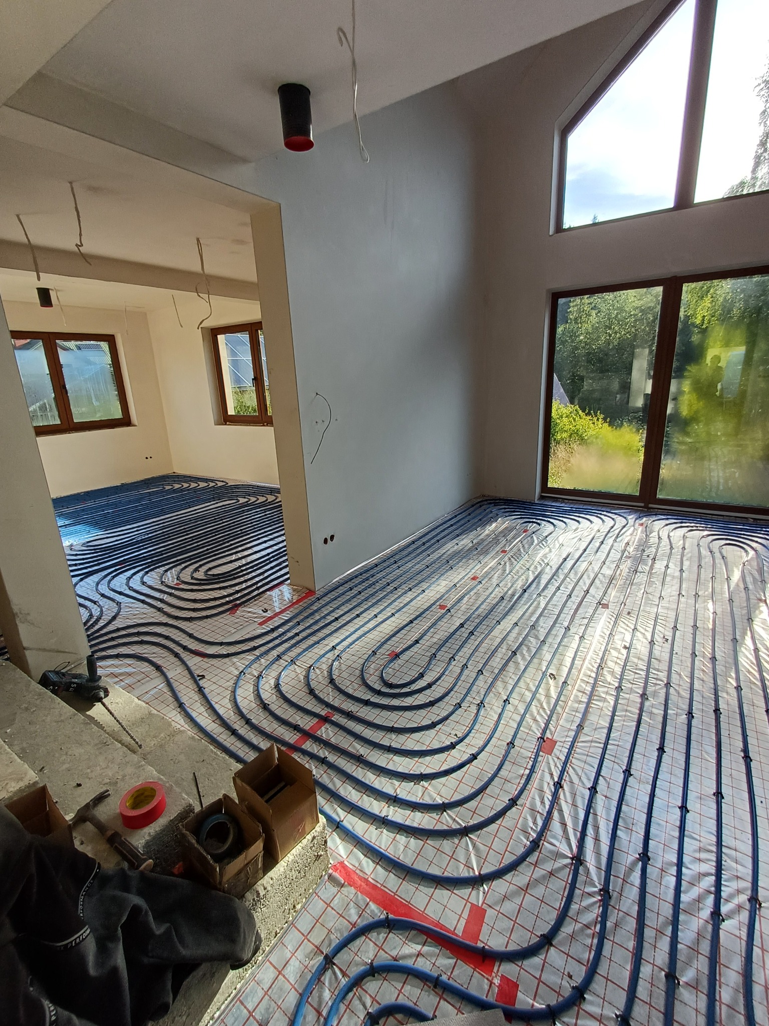 Układanie niebieskich rur ogrzewania podłogowego na folii z siatką w jasnym, przestronnym wnętrzu domu w stanie surowym, widoczne okna i narzędzia pracy.