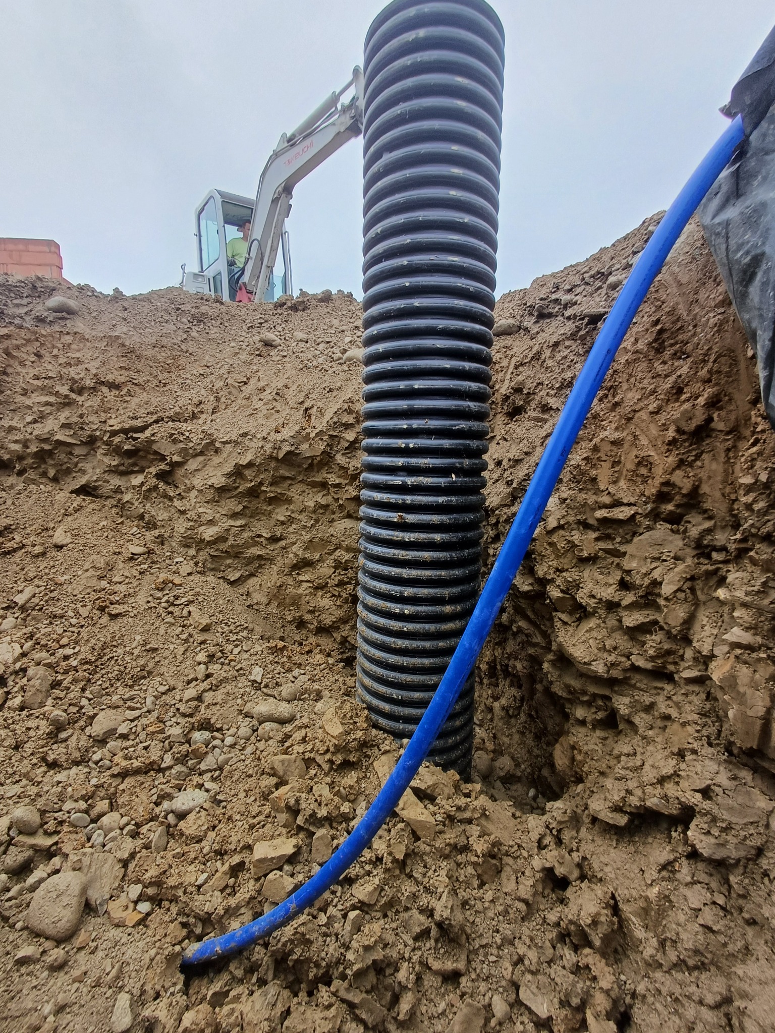 Czarne rury karbowane w wykopie ziemnym, obok błękitny przewód. W tle koparka i fragment ceglanej konstrukcji. Prace ziemne, instalacje podziemne.