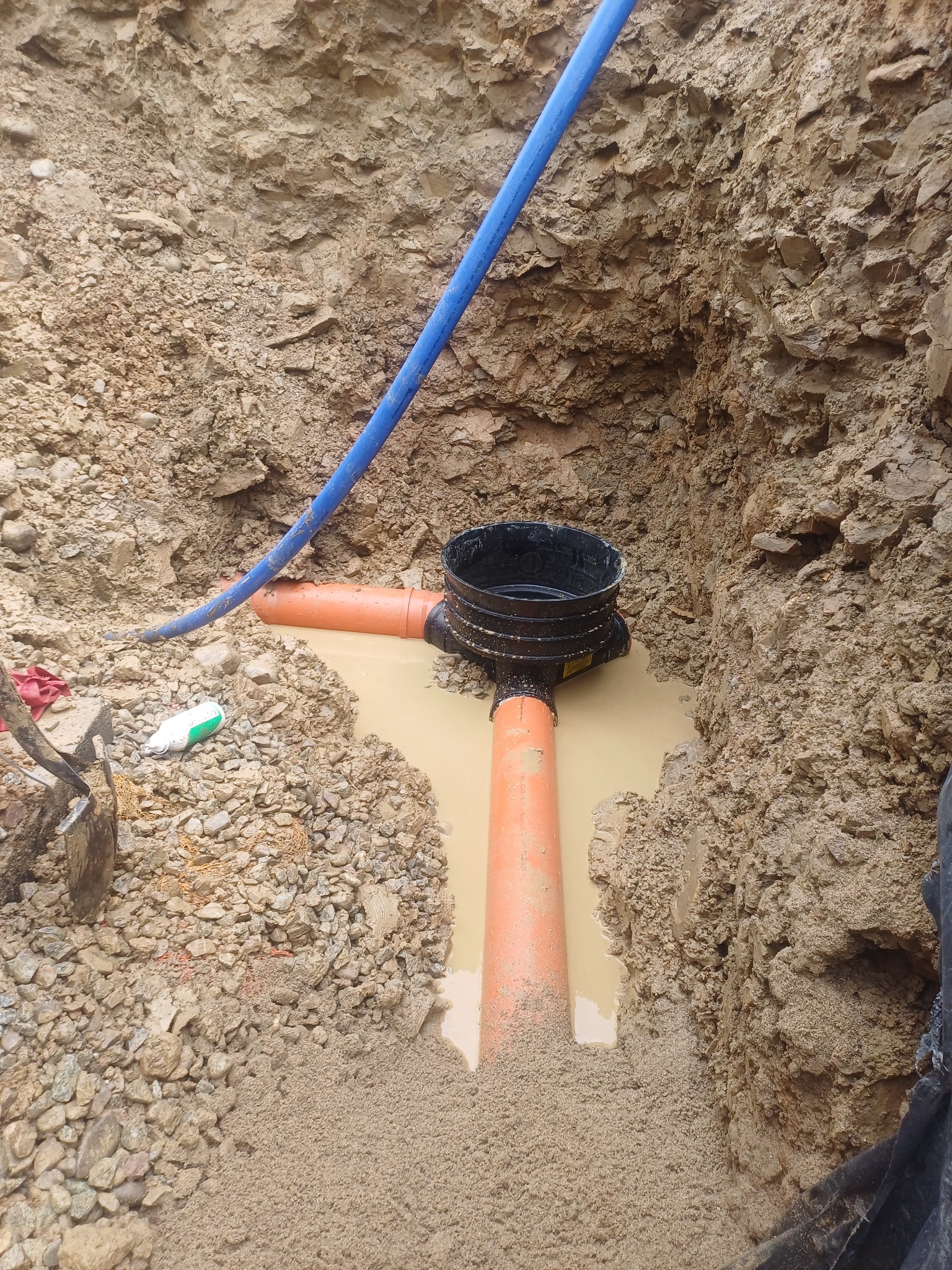 Wykop z pomarańczowymi rurami kanalizacyjnymi i czarnym zbiornikiem, częściowo zalany wodą. Widoczne narzędzia i niebieski przewód. Grunt skalisty.