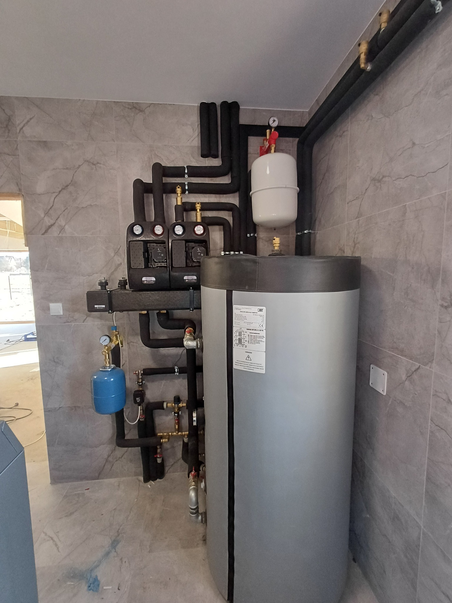 Zamknięta instalacja grzewcza z szarym zasobnikiem ciepłej wody, czarnymi rurami izolowanymi i elementami sterującymi na tle szarych płytek.