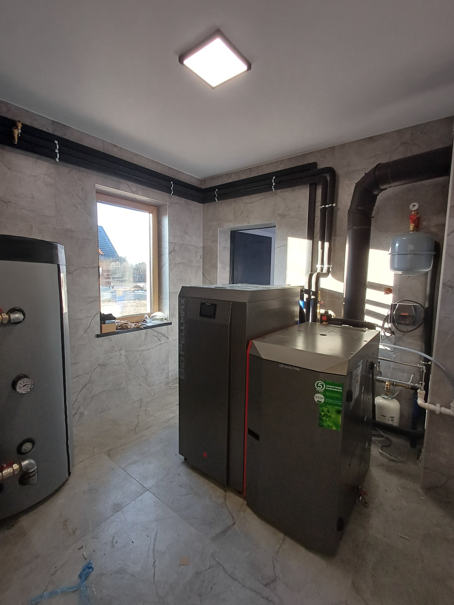 Nowoczesna kotłownia z piecem na pellet i zasobnikiem, rury grzewcze poprowadzone wzdłuż ścian wykończonych płytkami imitującymi marmur.