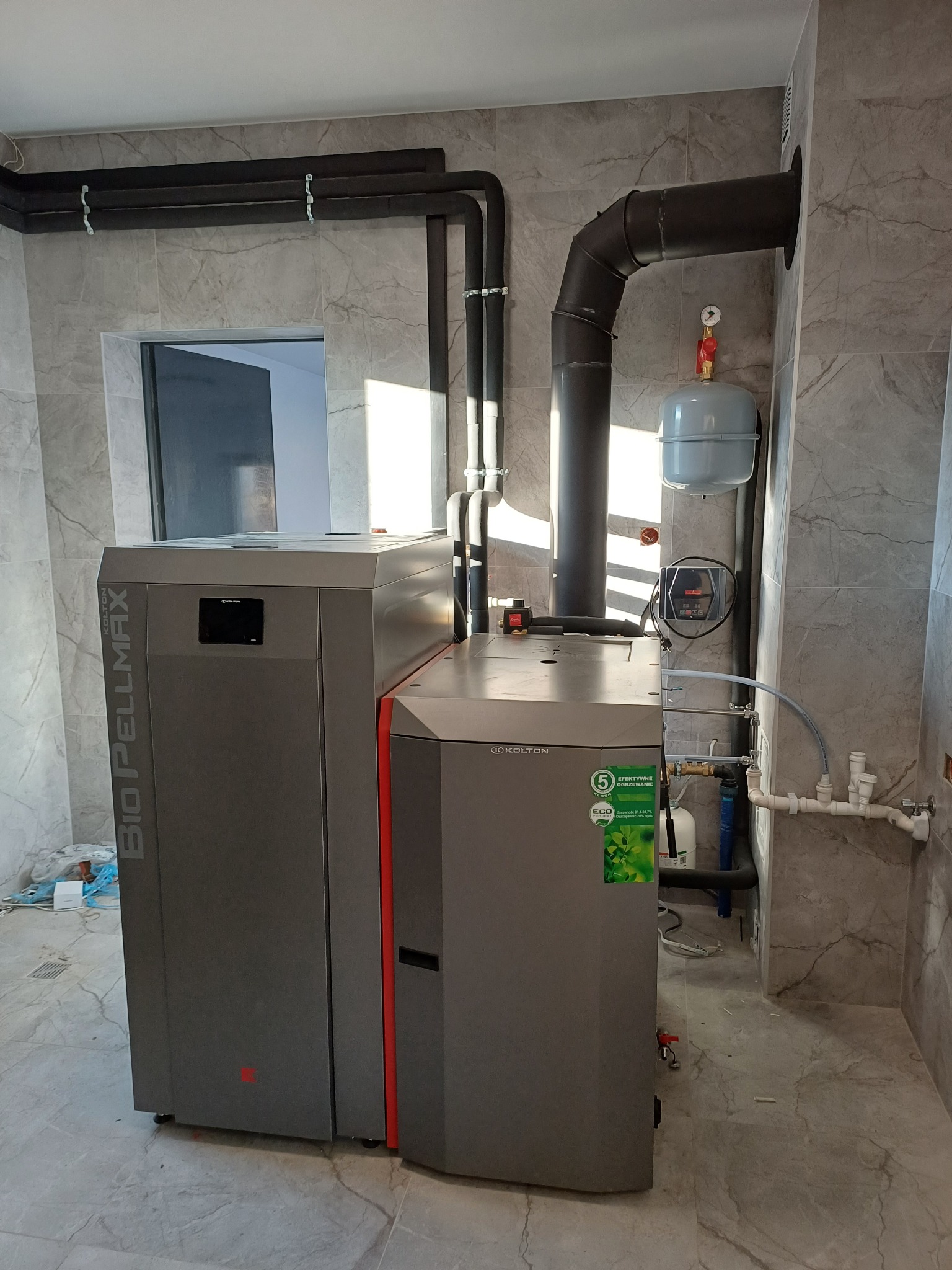 Szary piec na pellet Kolton Bio Pellmax z czerwoną wstawką, podłączony do instalacji grzewczej w pomieszczeniu wykończonym płytkami imitującymi marmur.