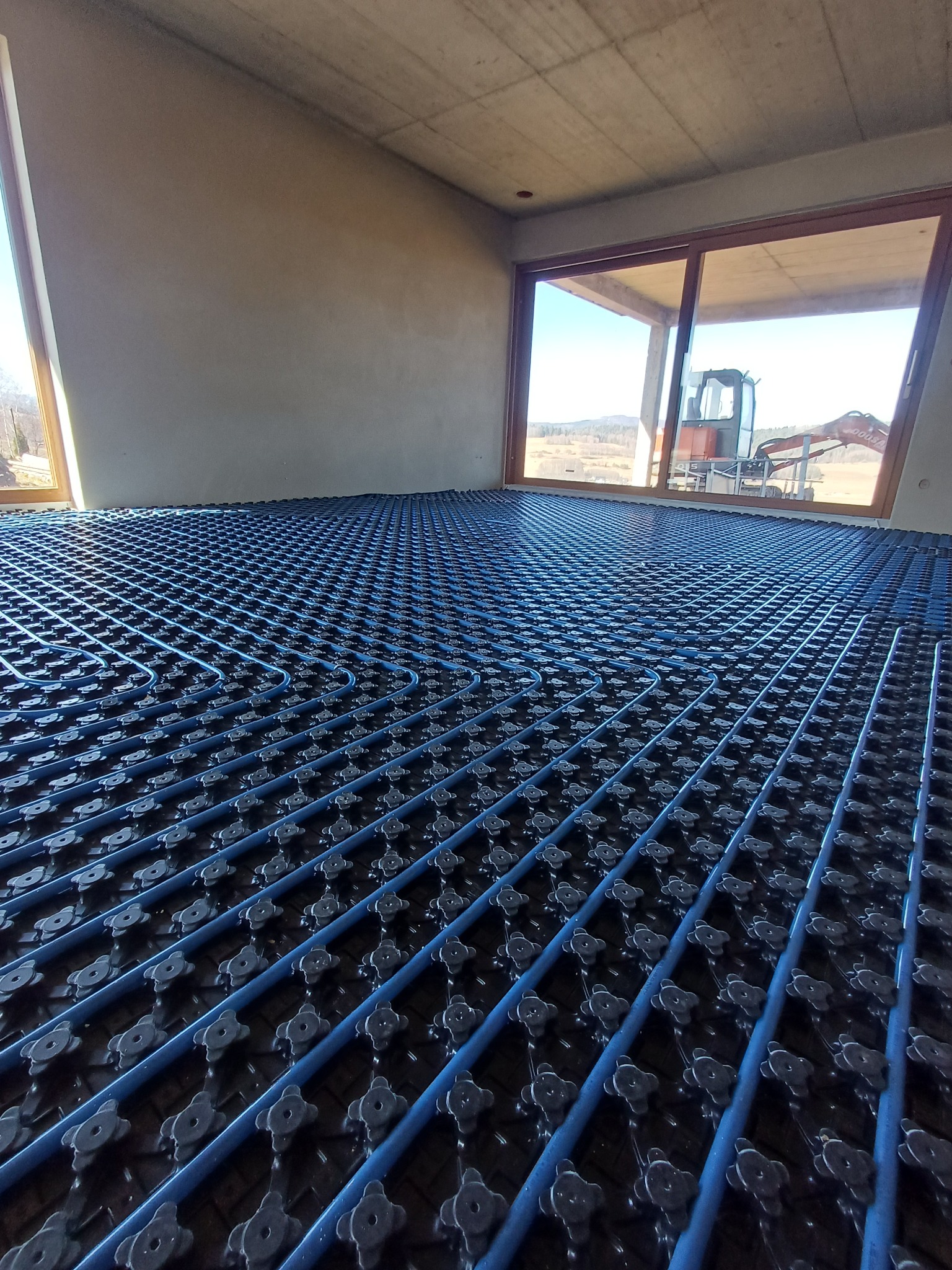 Niebieskie rury ogrzewania podłogowego ułożone w regularną siatkę na czarnej macie, widok z góry, w tle okno z widokiem na krajobraz i koparkę.