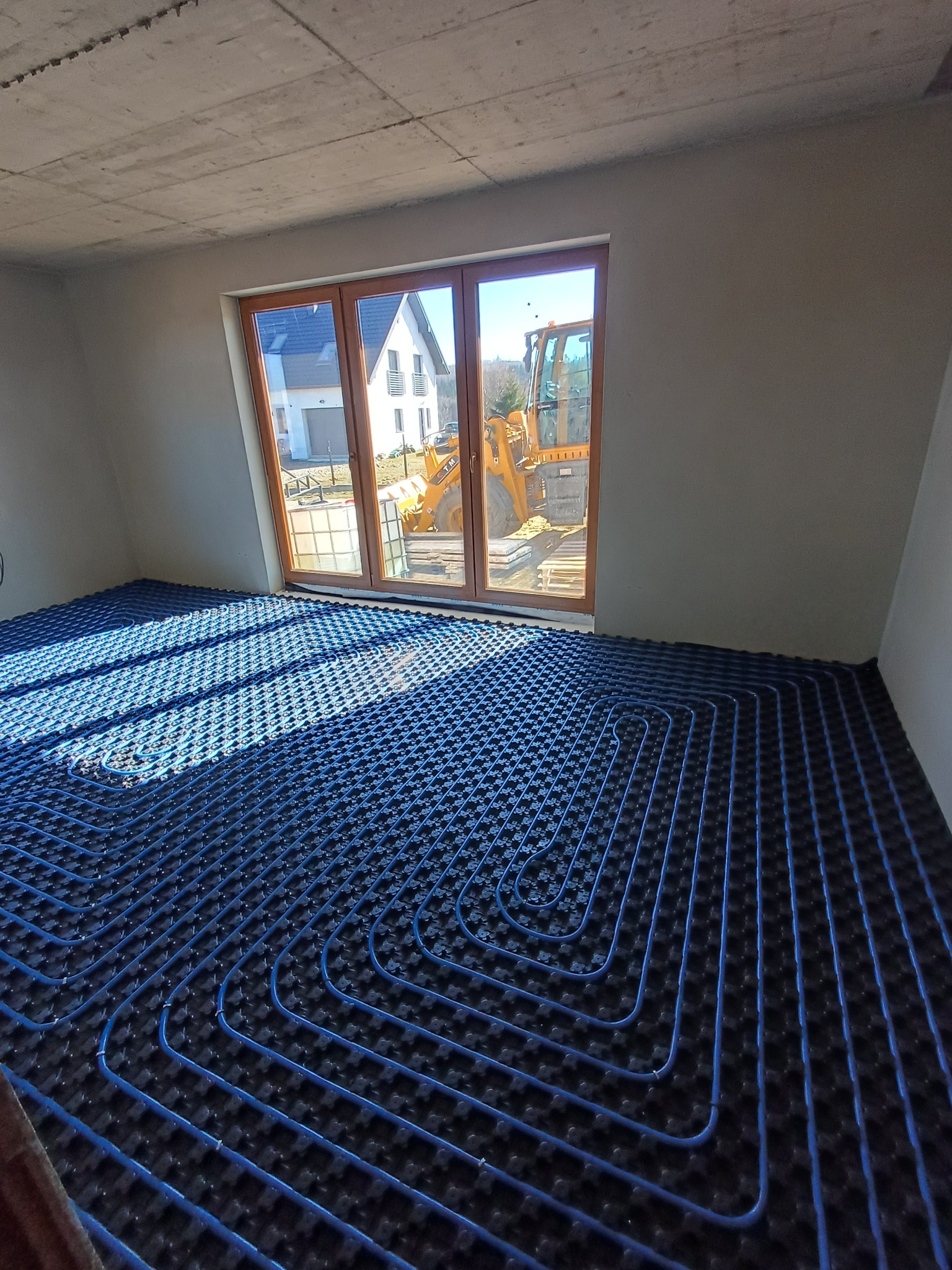 Niebieskie rury ogrzewania podłogowego ułożone w nowym budynku z widokiem na koparkę za oknem. Surowy betonowy sufit i szare ściany.