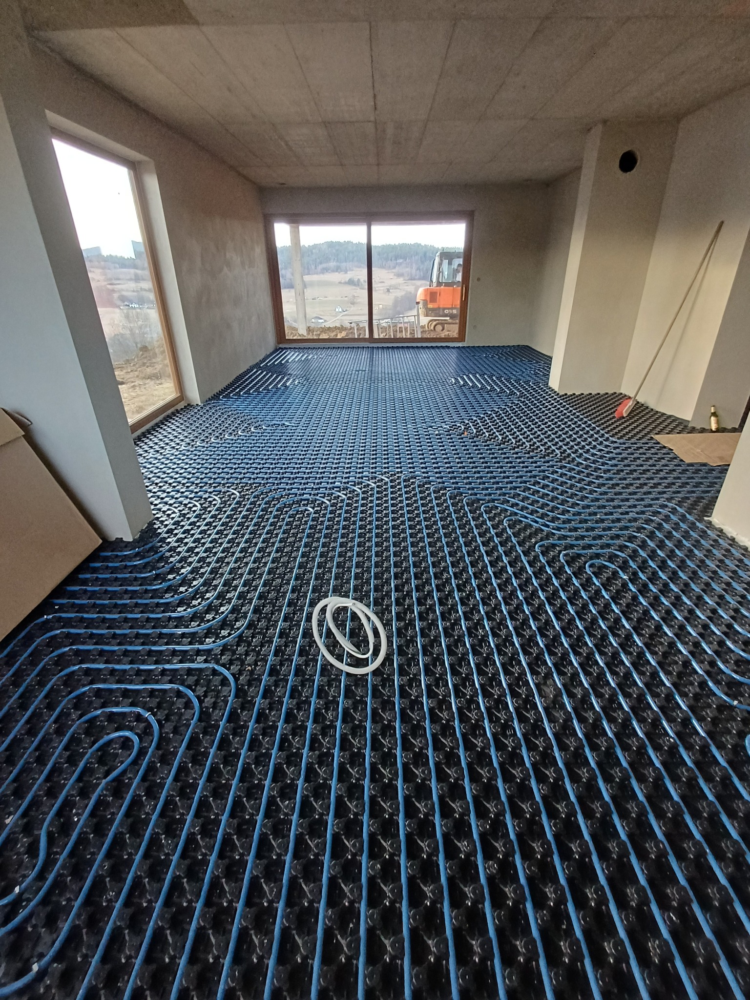 Niebieskie rury ogrzewania podłogowego ułożone na czarnej macie w surowym wnętrzu z oknami wychodzącymi na krajobraz. Widoczny sprzęt budowlany za oknem.