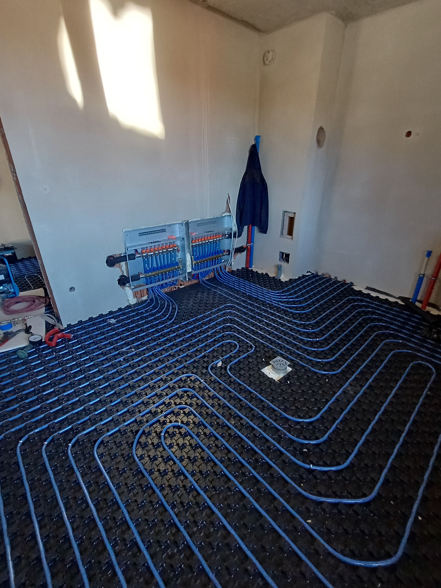 Niebieskie rury ogrzewania podłogowego ułożone na czarnej macie w surowym wnętrzu. Widoczny rozdzielacz i narzędzia. Ujęcie z góry, podkreślające regularny wzór instalacji.