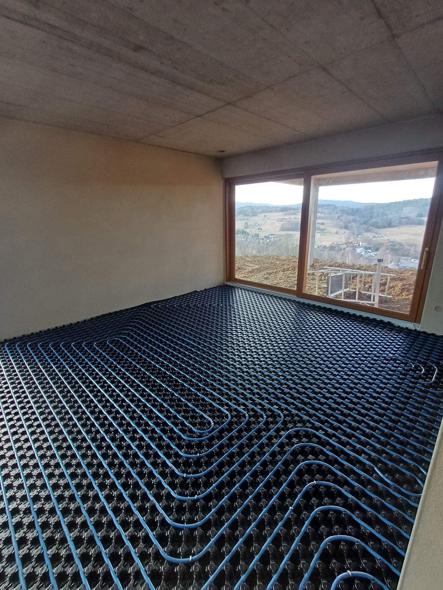 Niebieskie rury ogrzewania podłogowego ułożone na czarnej macie w surowym wnętrzu z widokiem na krajobraz przez okno. Instalacja przed wylaniem posadzki.