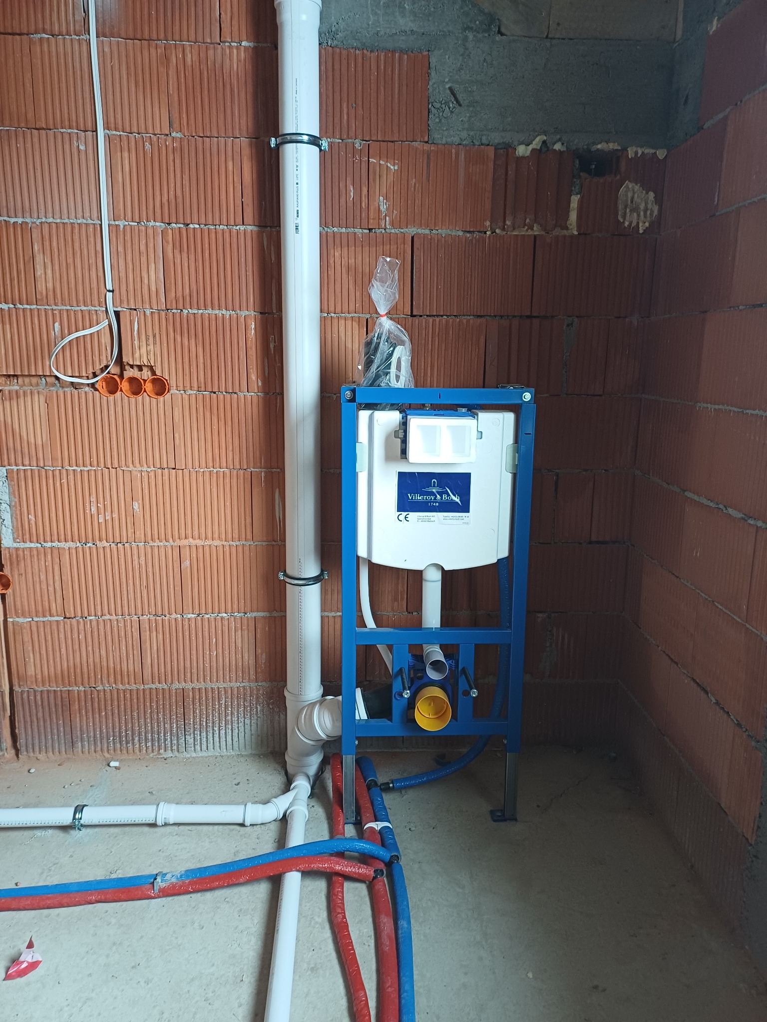 Instalacja podtynkowa WC Villeroy&Boch w surowym wnętrzu. Widoczne rury doprowadzające wodę (czerwone i niebieskie) oraz odpływ. Ściany z czerwonej cegły.