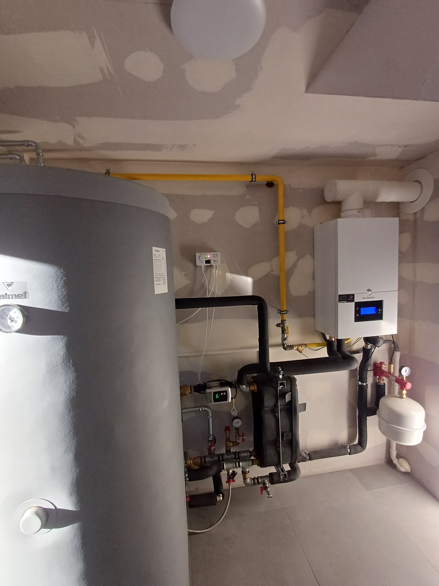 Nowoczesna kotłownia z szarym zasobnikiem ciepłej wody, białym piecem gazowym i żółtą instalacją gazową na tle surowych ścian w trakcie wykończenia.