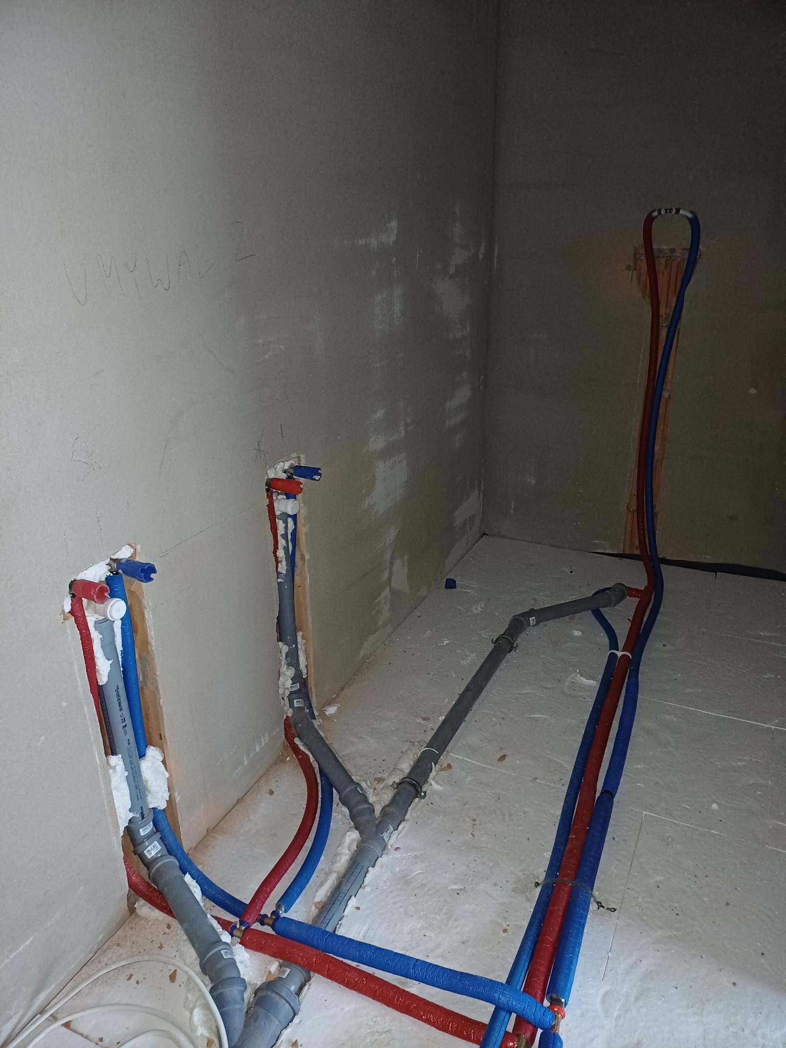 Fragment instalacji wodno-kanalizacyjnej w surowym wnętrzu, z rurami w kolorach czerwonym i niebieskim, oraz szarymi rurami odpływowymi. Na ścianie widoczny napis 'UMYWAL 2'.