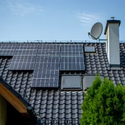 Czarne panele słoneczne na dachu domu z ciemnej dachówki, obok komina i anteny satelitarnej, widok z dołu na tle błękitnego nieba.