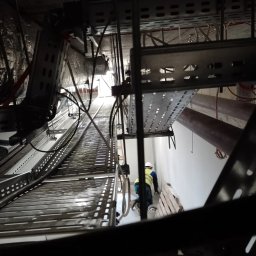 FEB ENERGIA - Montaż tras kablowych pod sufitem w nowym budynku w Warszawie, widok z perspektywy osoby stojącej na ziemi, pracownik w kasku i kamizelce odblaskowej przy ścianie.
