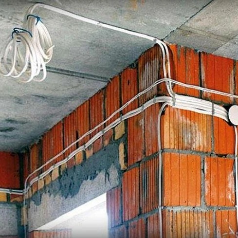 Białe przewody elektryczne prowadzone po ceglanej ścianie w surowym stanie, widoczne mocowania i pętle kabli zwisające z sufitu.