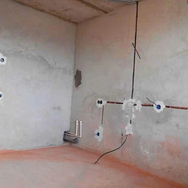 Surowy stan wnętrza z rozprowadzoną instalacją elektryczną w rurkach miedzianych i puszkami pod gniazdka, widoczne przewody i fragment instalacji hydraulicznej przy podłodze.