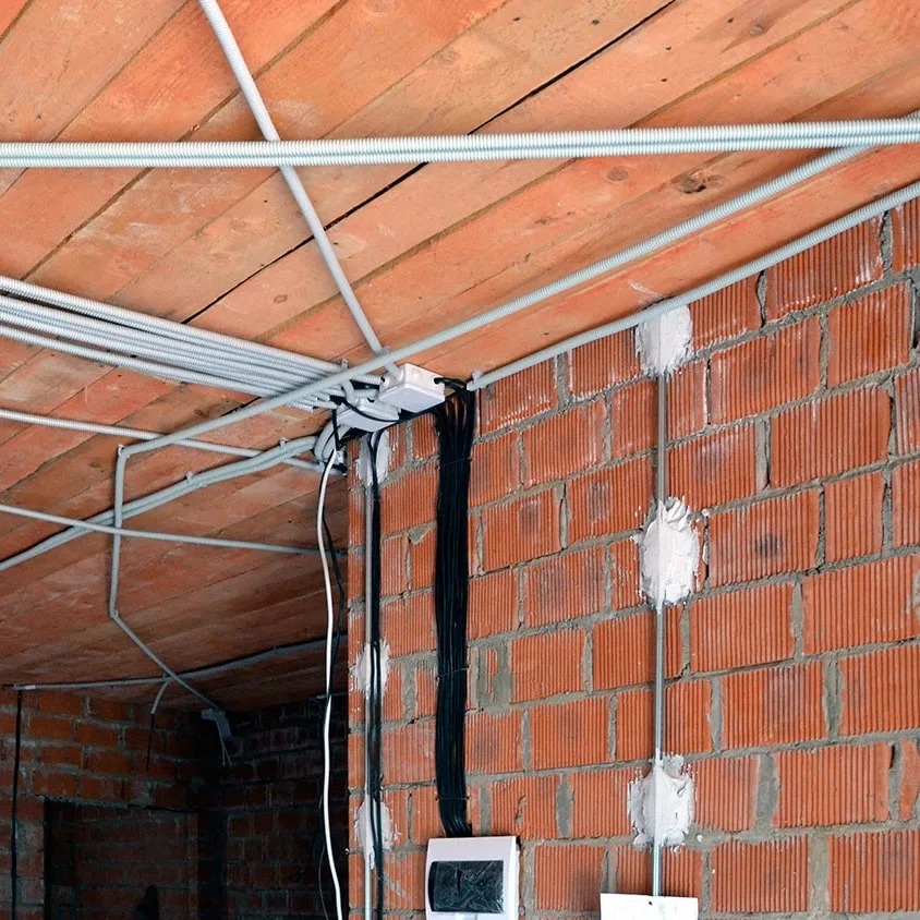 Instalacja elektryczna w budynku w trakcie budowy: kable w peszlach na ceglanym suficie i ścianie, puszki instalacyjne zalane gipsem.