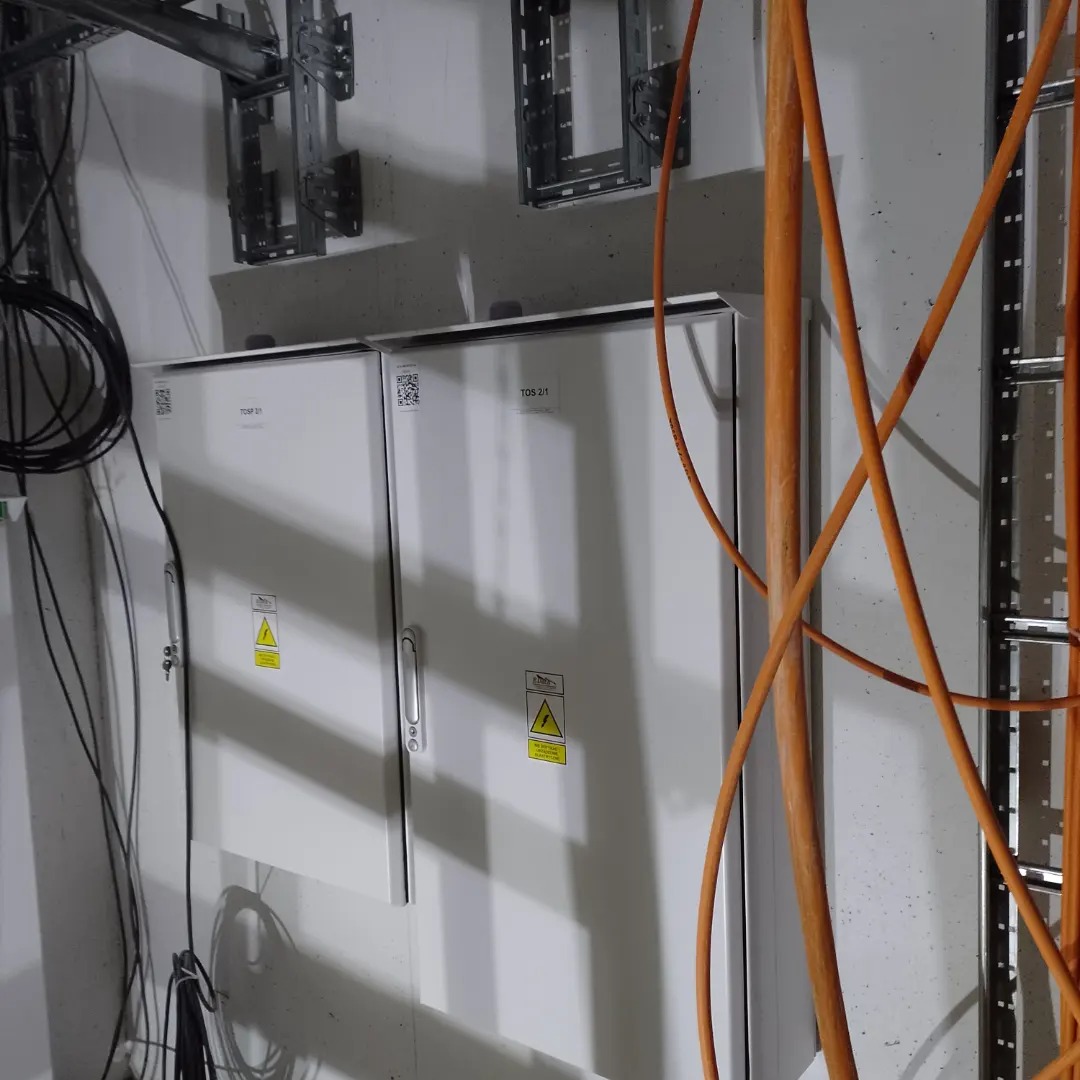 Dwa białe panele elektryczne z ostrzeżeniami, zamontowane na ścianie, z widocznymi przewodami i metalowymi wspornikami