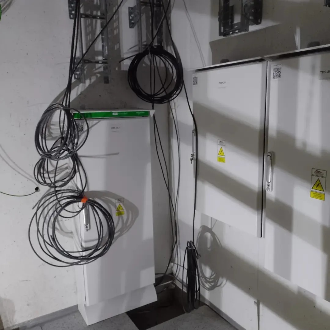 Dwa białe panele elektryczne z ostrzegawczymi naklejkami, zwisające kable i metalowe uchwyty na ścianie w pomieszczeniu technicznym.