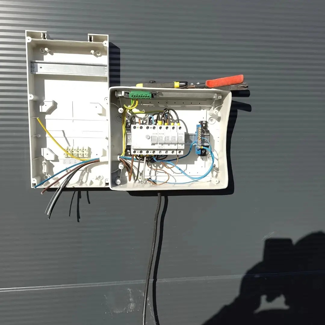 Otwarta skrzynka elektryczna z widocznymi przewodami, bezpiecznikami i narzędziami, zamontowana na szarej ścianie z poziomymi przetłoczeniami.