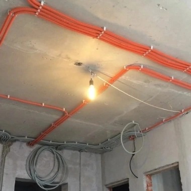 Pomarańczowe rury instalacyjne przymocowane klamrami do surowego, betonowego sufitu. Widoczna wisząca żarówka i szare kable w ścianie w trakcie montażu.