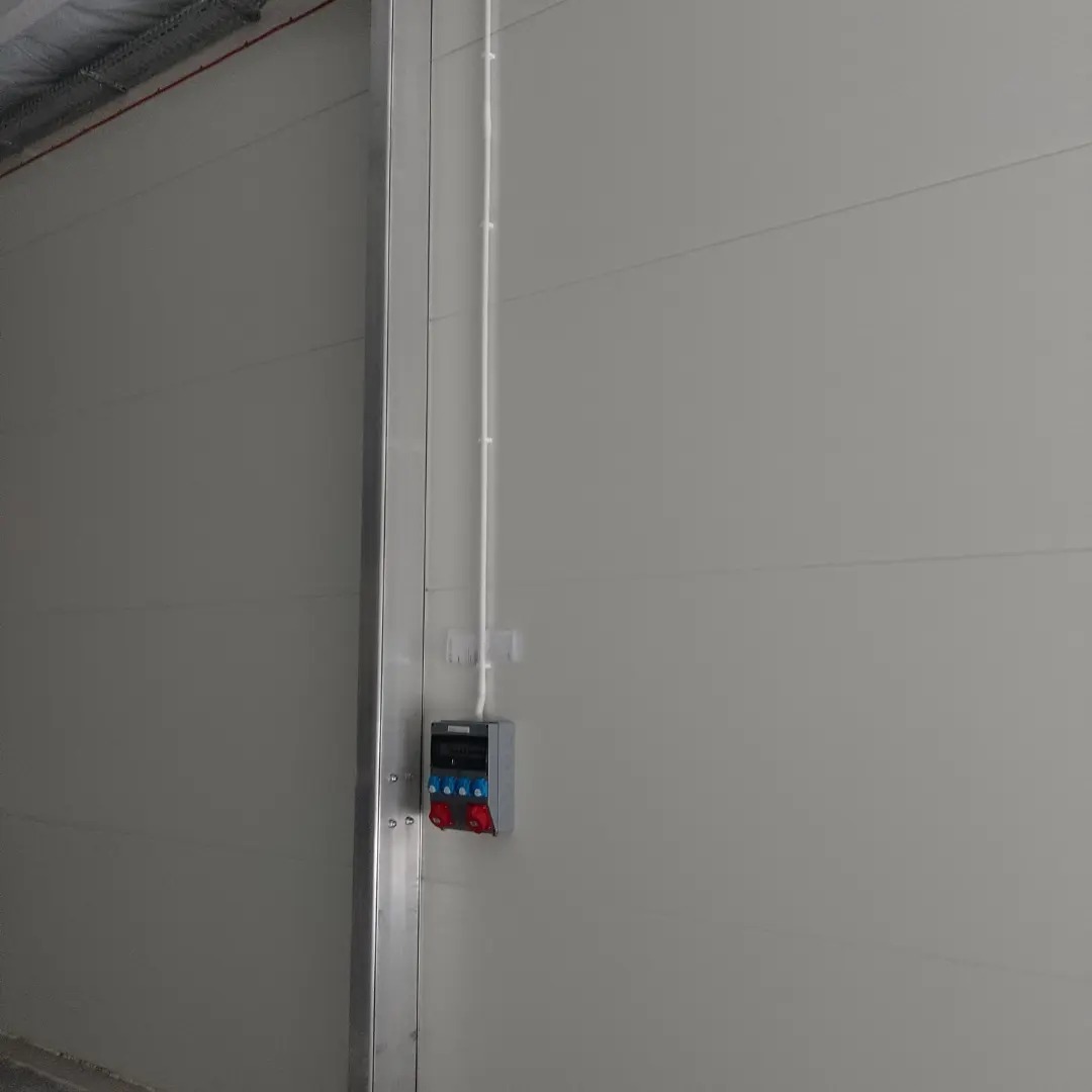 Szara ściana hali z zamontowaną szarą skrzynką elektryczną z czerwono-niebieskimi gniazdami i białą listwą kablową.