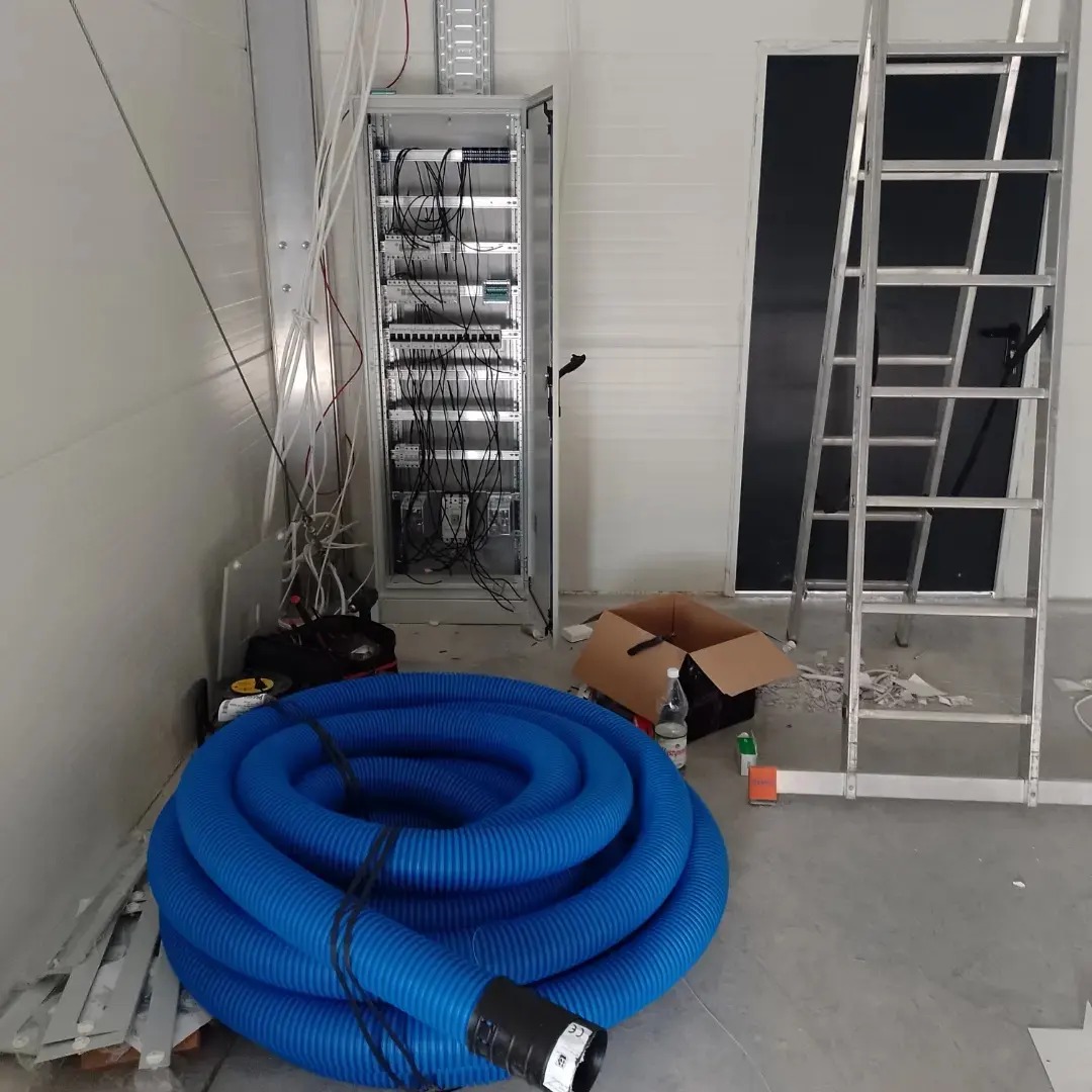 Szafa serwerowa z podłączonymi kablami, zwój niebieskiego węża wentylacyjnego i aluminiowa drabina w surowym wnętrzu z panelami ściennymi