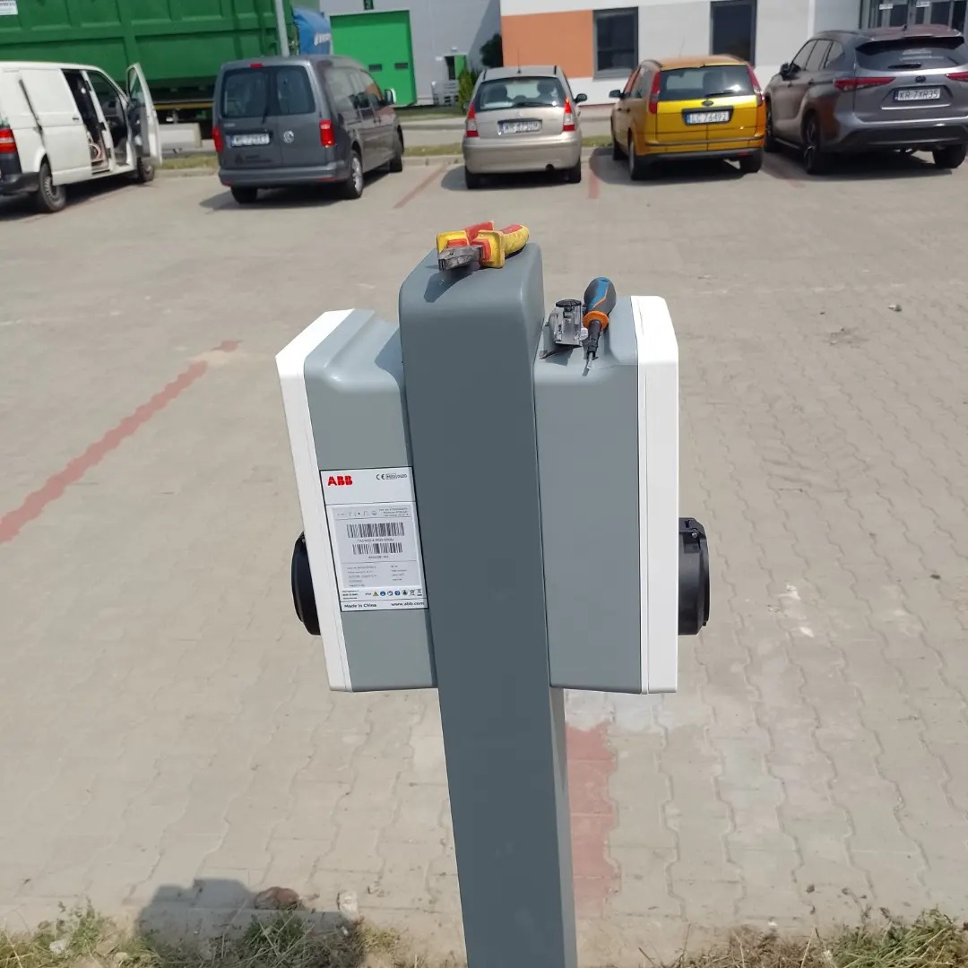 Szaro-biała stacja ładowania pojazdów elektrycznych marki ABB na tle parkingu z samochodami, z narzędziami (kombinerki i śrubokręt) leżącymi na górze obudowy.