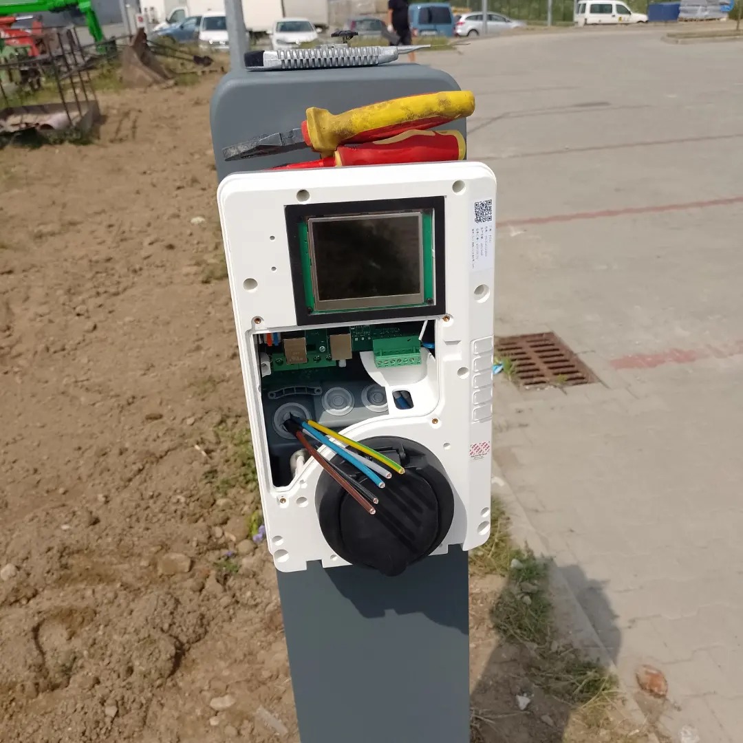 Otwarta obudowa stacji ładowania pojazdów elektrycznych z widocznymi przewodami, umieszczona na szarym słupku, z kombinerkami leżącymi na górze.
