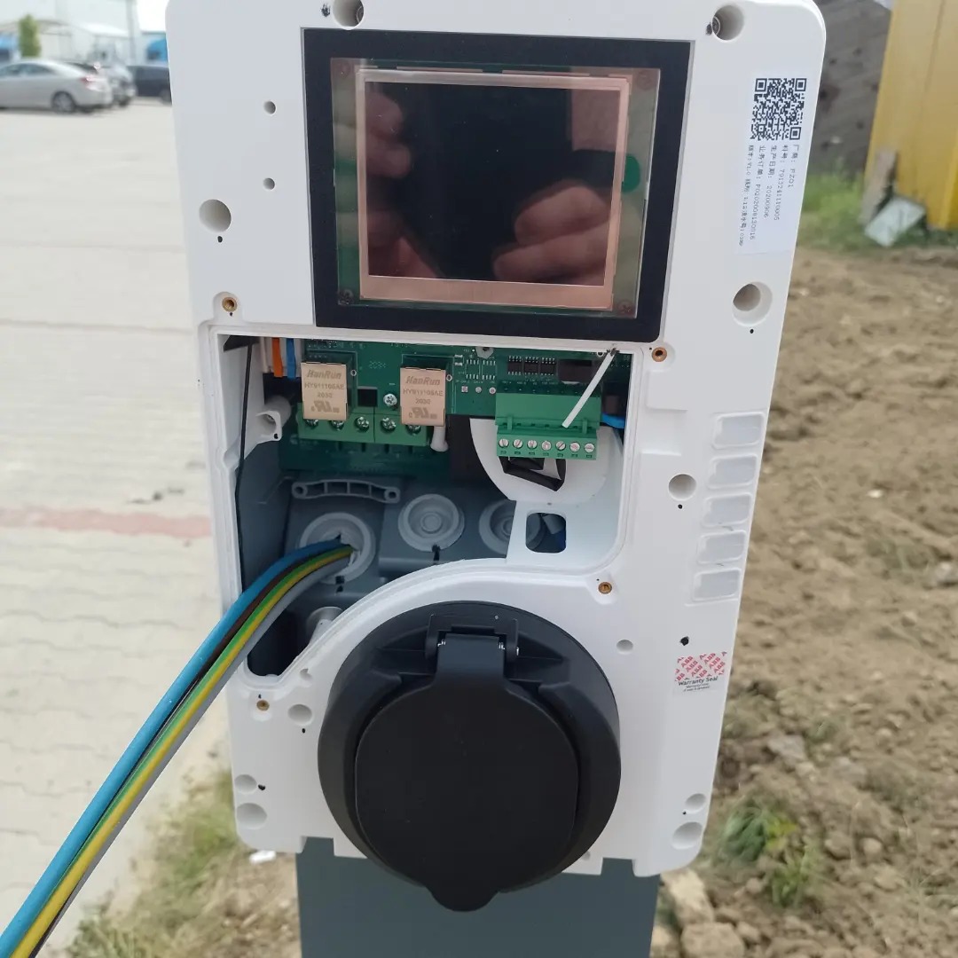 Otwarta obudowa stacji ładowania pojazdów elektrycznych z widocznymi kablami, elektroniką i ekranem, zainstalowana na zewnątrz.