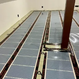 Montaż mat grzewczych marki BRAAN na podłodze w pomieszczeniu z drewnianym słupem nośnym, widoczne czarne taśmy z napisem BRAAN.