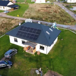 Widok z drona na dom jednorodzinny z ciemną dachówką i panelami słonecznymi na dachu, otoczony zielonym trawnikiem, z dwoma samochodami na podjeździe i grupką osób w pobliżu.