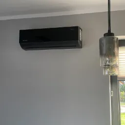 Czarna jednostka klimatyzacyjna Rotenso Elis zamontowana na szarej ścianie w nowoczesnym wnętrzu, obok widoczna lampa wisząca i okno z żaluzjami.