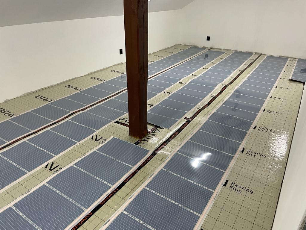 Montaż folii grzewczej na podłodze w pomieszczeniu z drewnianym słupem nośnym, widoczne arkusze folii grzewczej z nadrukiem 'Heating Film' i logo producenta.