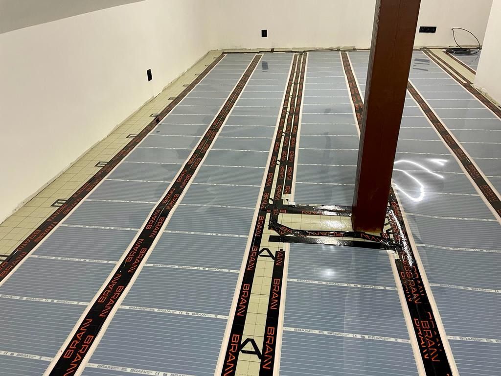 Montaż mat grzewczych marki BRAAN na podłodze w pomieszczeniu z drewnianym słupem nośnym, widoczne czarne taśmy z napisem BRAAN.