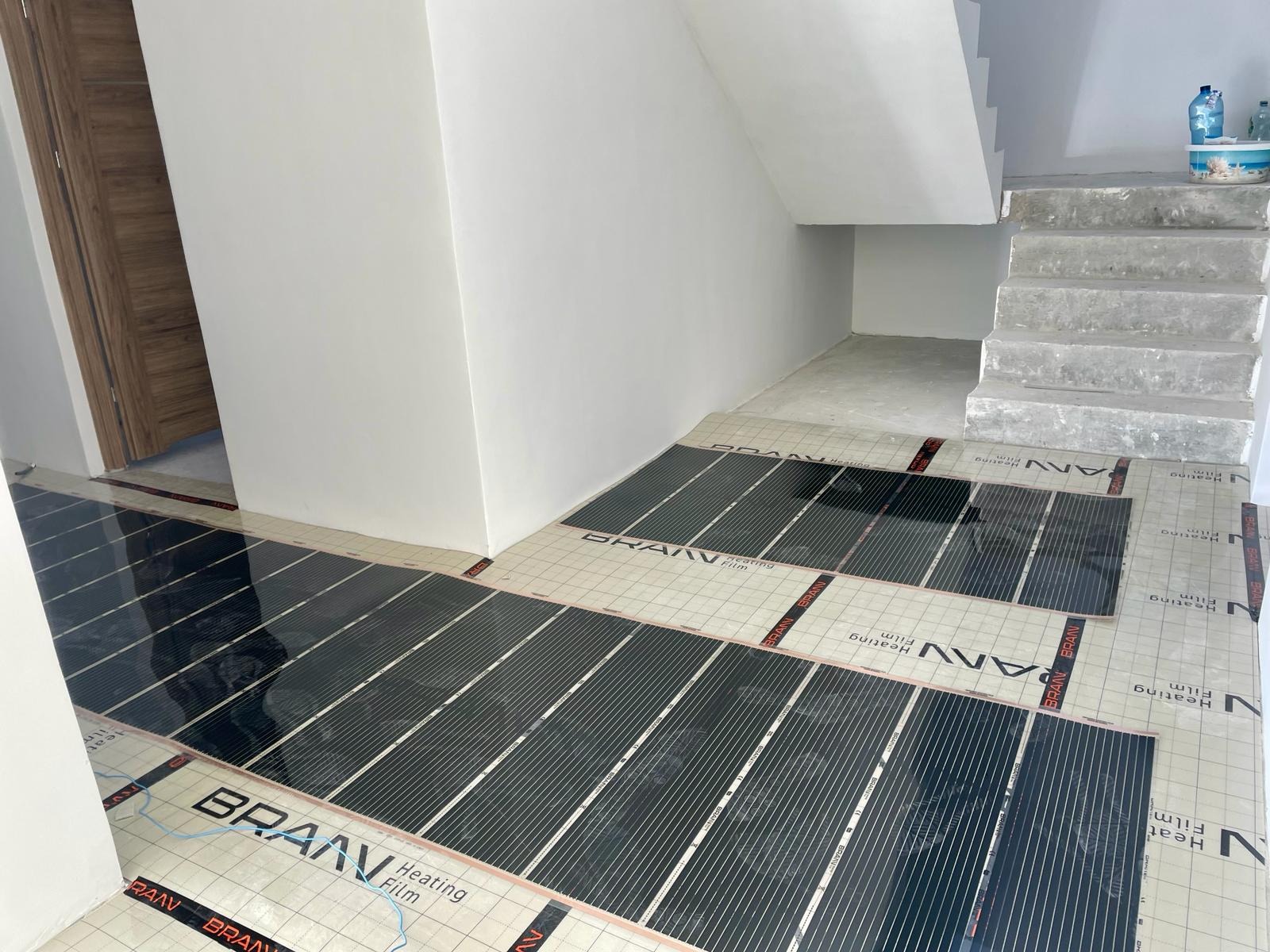 Rozłożone maty grzewcze marki BRAAN Heating Film na podłodze w jasnym pomieszczeniu z surowymi schodami betonowymi, przygotowane do dalszych prac wykończeniowych.