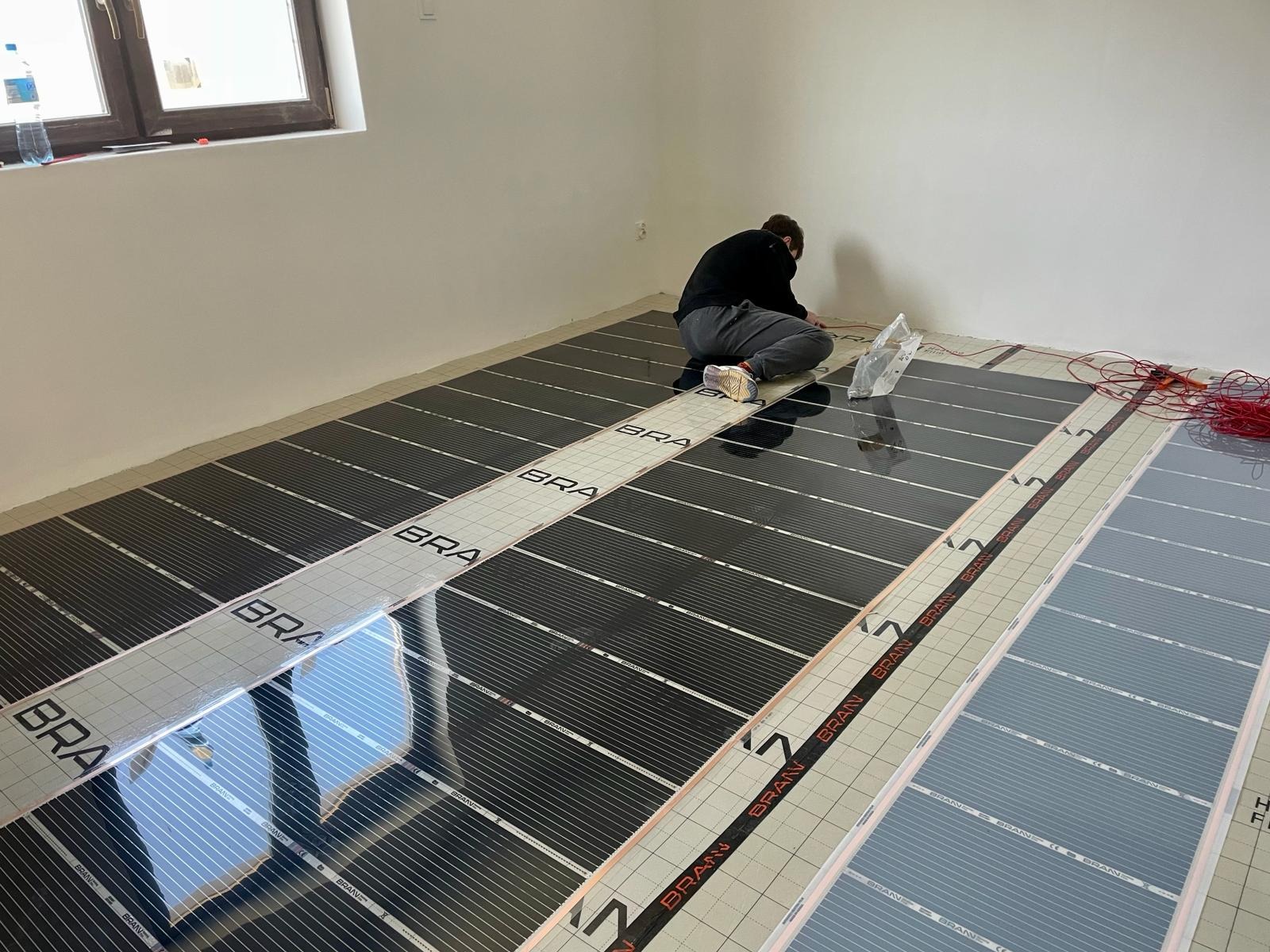 Instalacja elektrycznych mat grzewczych podłogowych w nowym budynku; widoczne czarne maty z napisem 'BRAN' układane na podłodze, instalator łączy kable