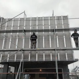 Montaż paneli elewacyjnych z betonu architektonicznego na rusztowaniu, widoczni dwaj pracownicy w odzieży roboczej, szare niebo w tle.