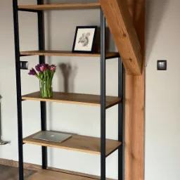 Regał z drewnianymi półkami i czarną metalową ramą, na półkach wazon z tulipanami, ramka ze szkicem wiewiórki i laptop, na tle drewniany element konstrukcyjny i szara ściana.